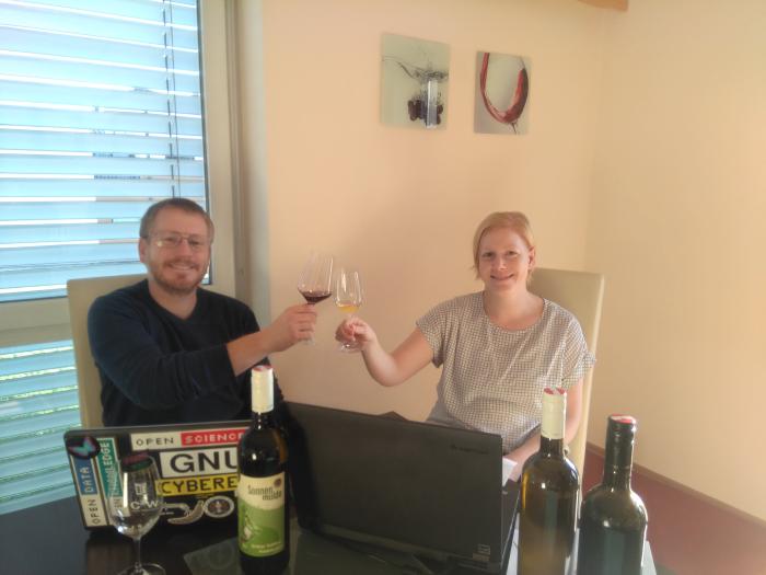 Kathrin and Andreas are sitting at a table with two laptops on it. They toast each other with two wine glasses. There are a few wine bottles on the table.
