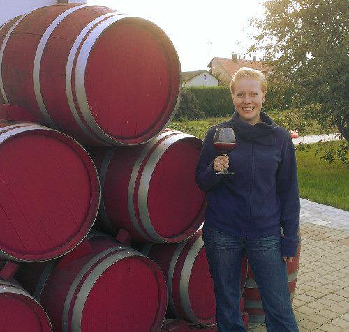 Kathrin steht mit einem Glas Traubensaft neben einem Stapel rot gestrichener Barriquefässer die als Dekoration im Garten dienen.