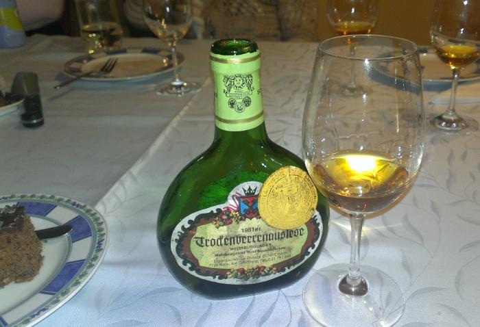 Eine geöffnete Flasche TBA mit eingeschenktem Glas auf einem Tisch. Daneben ein Teller mit Schokoladentorte.