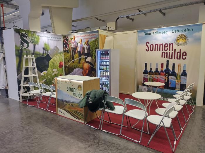Ein Messestand mit Teppich, Tischen und Sesseln. Am Gang ein Pult mit aufgedrucktem Bild und Logo. Im Hintergrund Wände mit abgebildeten Weinflaschen und Menschen.