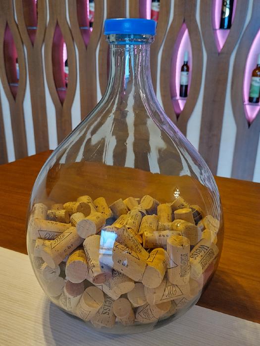 Auf einem Tisch steht ein Glasballon, eine etwa 20 L große ballonförmige Glaschflasche. Sie ist etwa bis zur Hälfte des bauchigen Teils mit verschiedenen Weinkorken gefüllt. Im Hintergrund sieht man eine Wand mit Nischen in denen Weinflaschen präsentiert werden.