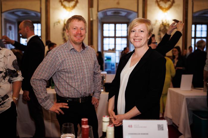 Andreas and Kathrin stand behind their wine bottles and smile at the camera