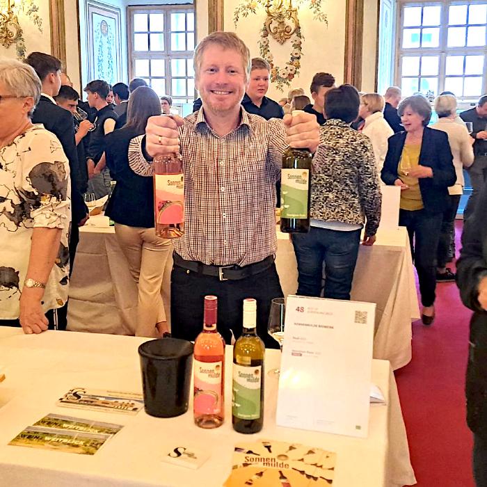 Andreas stands grinning behind a table with a white tablecloth and holds a bottle each of Rosé and Sauvignon Blanc into the camera