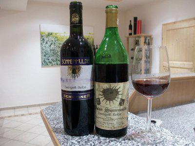 Two bottles of wine in completely different styles. The obviously older bottle is opened and in a glass next to it there has been some red wine poured in.