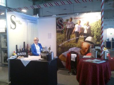 View of the exhibition stand. In the foreground counters with wine bottles for presentation. On the back wall large posters with pictures of people and wine bottles.