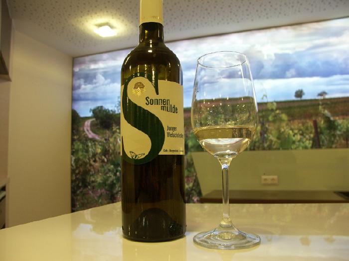 A bottle and a glass with some wine stand on a white counter. In the background there is an illuminated picture of a vineyard.