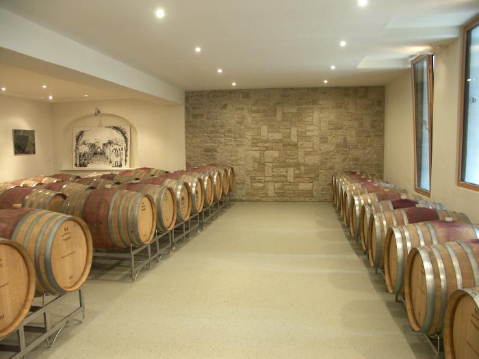 Cellar with central corridor and stone wall at its end. Barrique barrels lined up on metal racks to the left and right of the aisle.