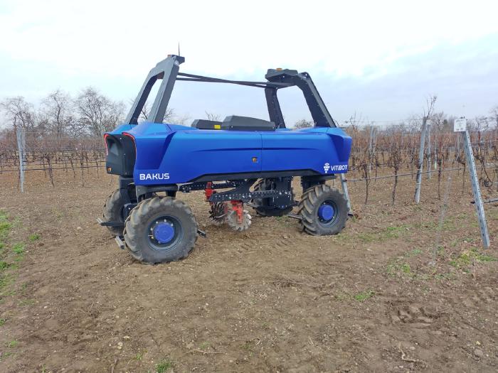 Vineyard robot from the side. You can see a narrow chassis with 360 degree rotating tractor tires at both ends. Two frames connect the body to a mirrored, identical part on the opposite side. The device can thereby take a row of vines in the middle and carry out over-the-row work. The robot is about 5m long and 2.2m high with a width of a good 1.5m.