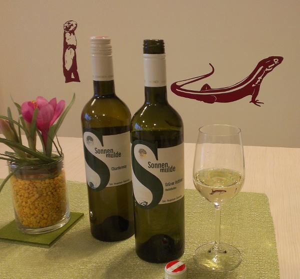 Auf einem Tisch stehen zwei Weinflaschen, eine davon geöffnet. Daneben ein leicht gefülltes Glas und eine Vase mit Dekoration. Im Hintergrund Grafiken mit einem Ziesel und einer Eidechse an der Wand.