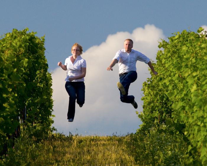 Kathrin and Andreas run down between rows of vines and jump up into the air with their feet stretched out.